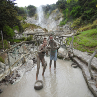 A mud-covered couple poses in a thermal pool surrounded by vegetation and mountains.