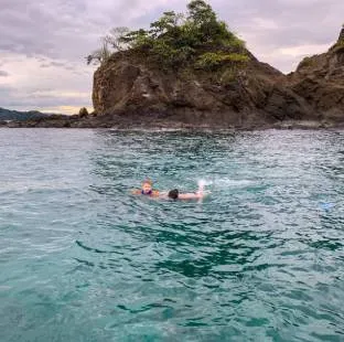 Two people swim in calm waters in front of a rock formation covered in vegetation.