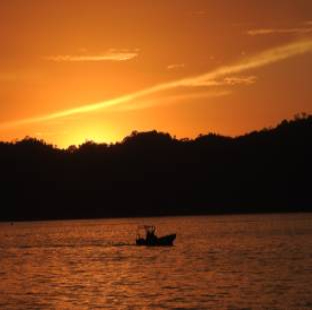 A small boat sails on calm water at sunset, with a dark silhouette of a hilly shoreline in the background and an orange sky overhead.