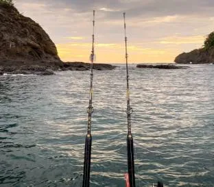 Two fishing rods extend over calm ocean waters at sunset near rocky coastal cliffs.