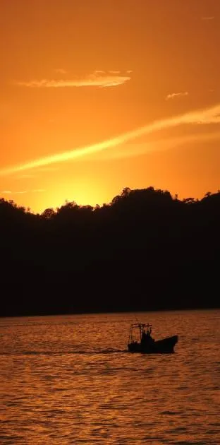 A small boat sails on calm water at sunset, with a dark silhouette of a hilly shoreline in the background and an orange sky overhead.