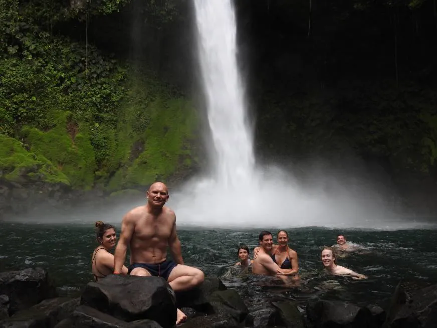 Two men sit at the base of a waterfall cascading over dark rocks into a natural pool.