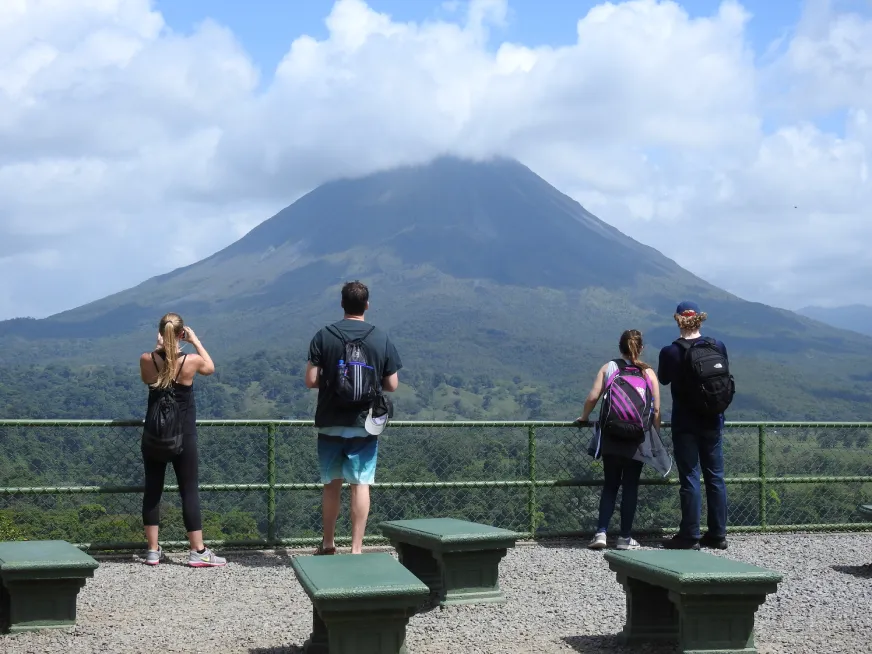 Three tourists stand on a viewing platform overlooking Arenal Volcano surrounded by green vegetation.