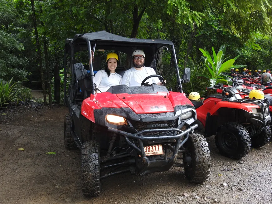 A couple wearing helmets drives a red off-road vehicle on a trail surrounded by vegetation.