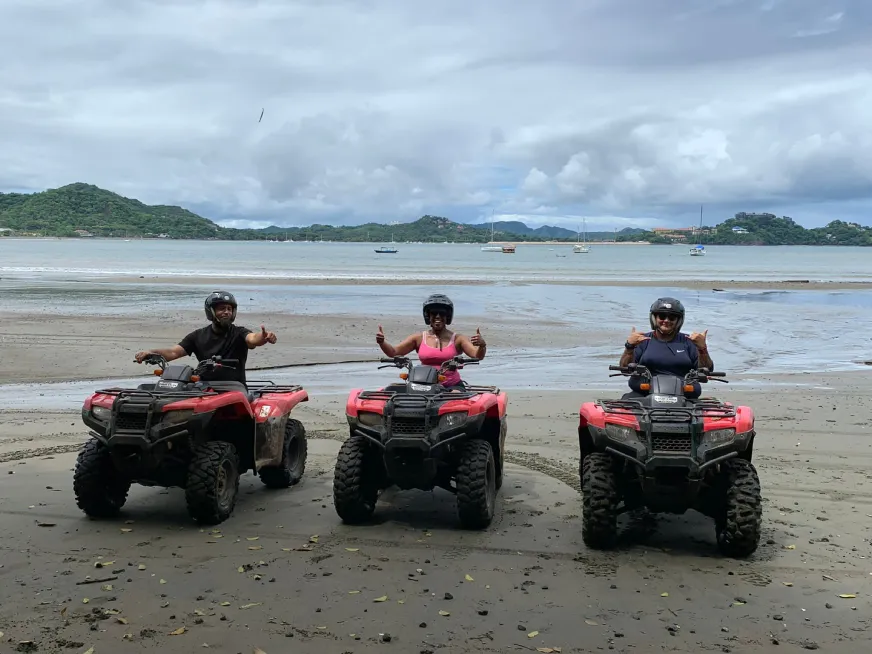 Three people wearing helmets ride red ATVs on a trail in front of green, wooded hills.