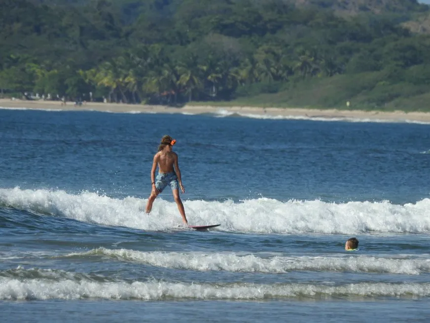 A young man surfs on a wave while another child swims nearby in the tropical sea.