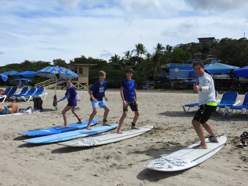 An instructor and three children practice balancing on surfboards on a sunny beach.