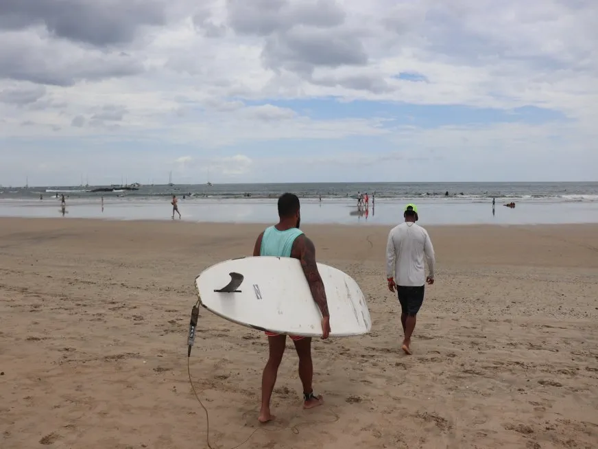 Two men walk along the beach, one of them carrying a white surfboard.