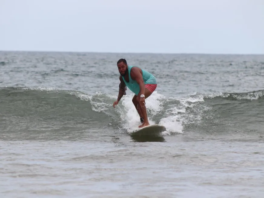 A man wearing a turquoise tank top and patterned swim trunks surfs on a wave in the sea.