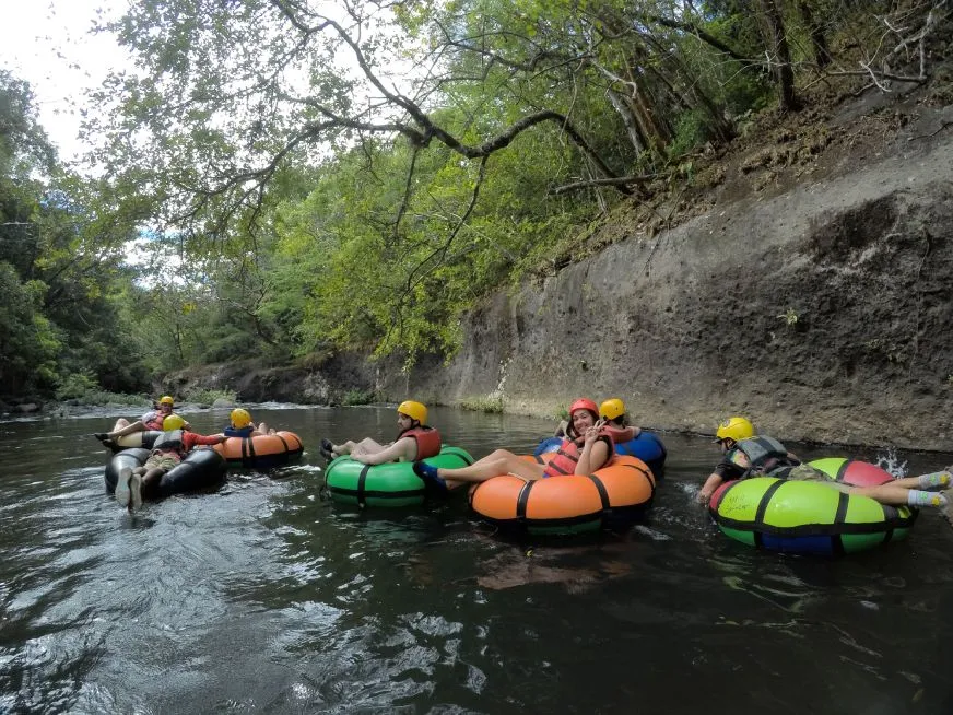 Group of people wearing helmets and life jackets floating on colorful inner tubes down a river surrounded by vegetation.