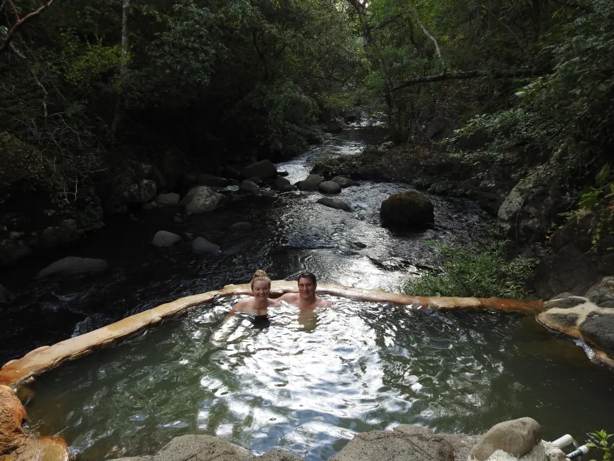 A smiling couple enjoys a natural thermal pool surrounded by vegetation and a nearby river.