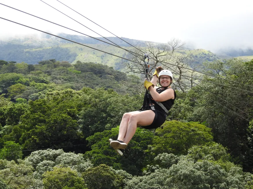 A woman zip lines through a dense green forest canopy with a panoramic mountain view.