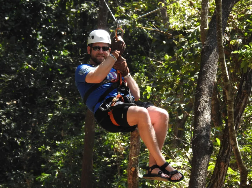 A woman wearing a helmet and harness zip lines through a dense green tropical forest.