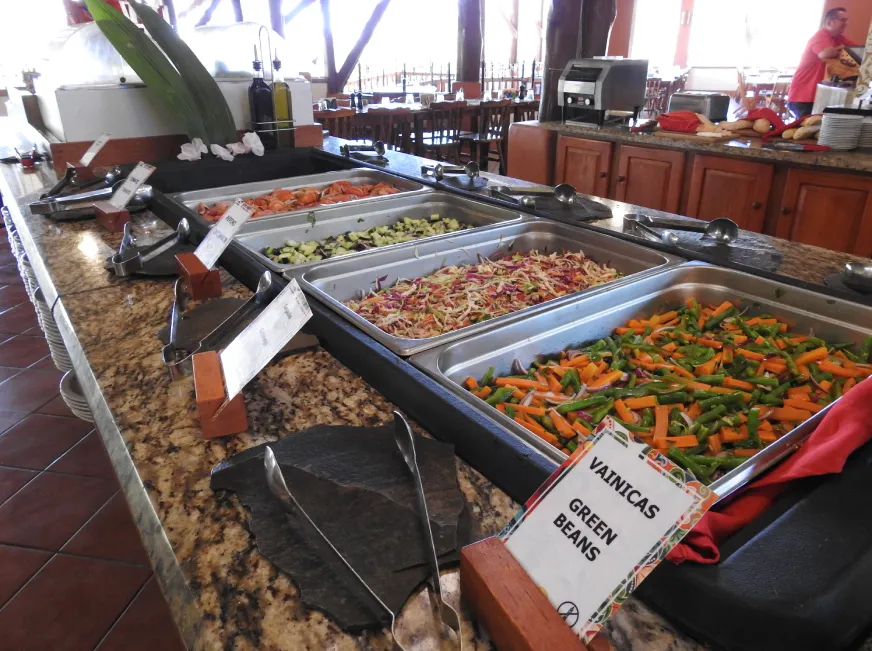 A tourist photographs a colorful spread of tropical fruits on a buffet table indoors.