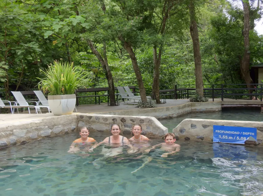 A group of people relax in a natural thermal pool surrounded by tropical vegetation.