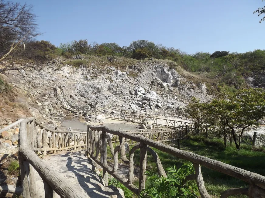 A wooden footbridge crosses a barren volcanic landscape with rocky grey terrain under a blue sky.