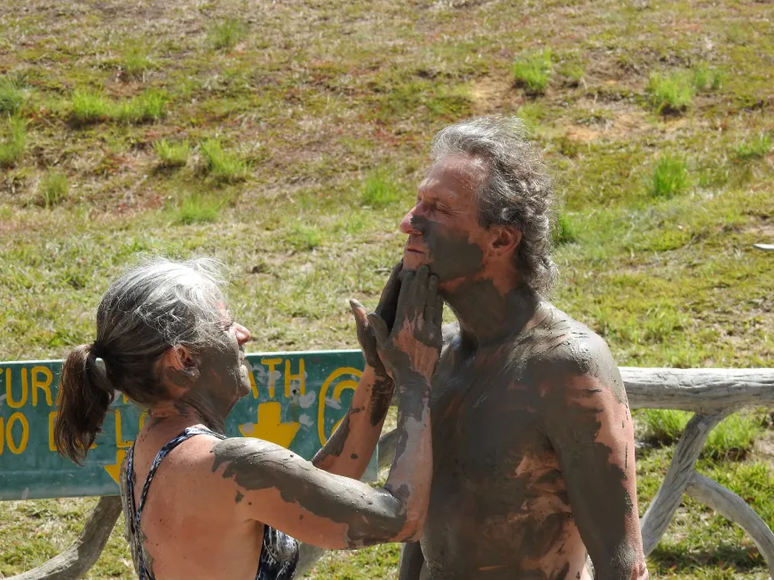 Two people covered in volcanic mud stand on a wooden bridge surrounded by tropical forest.