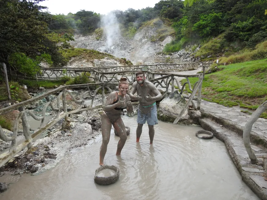 Two tourists stand on a wooden walkway surrounded by volcanic steam vents and grey rocky terrain.
