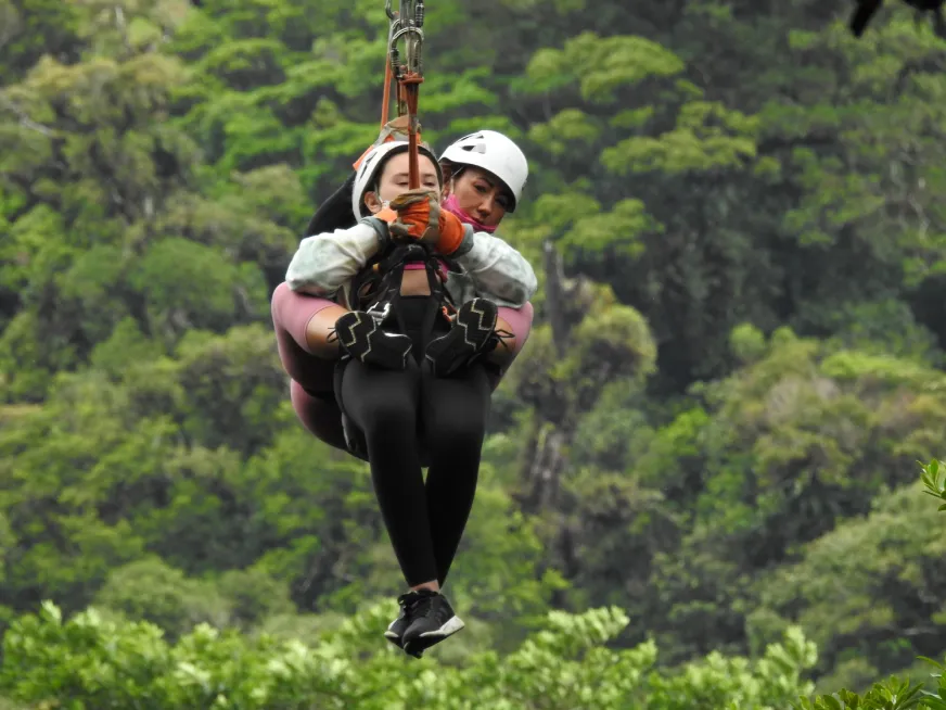 Two people wearing helmets and harnesses glide together down a zip line over a green forest.