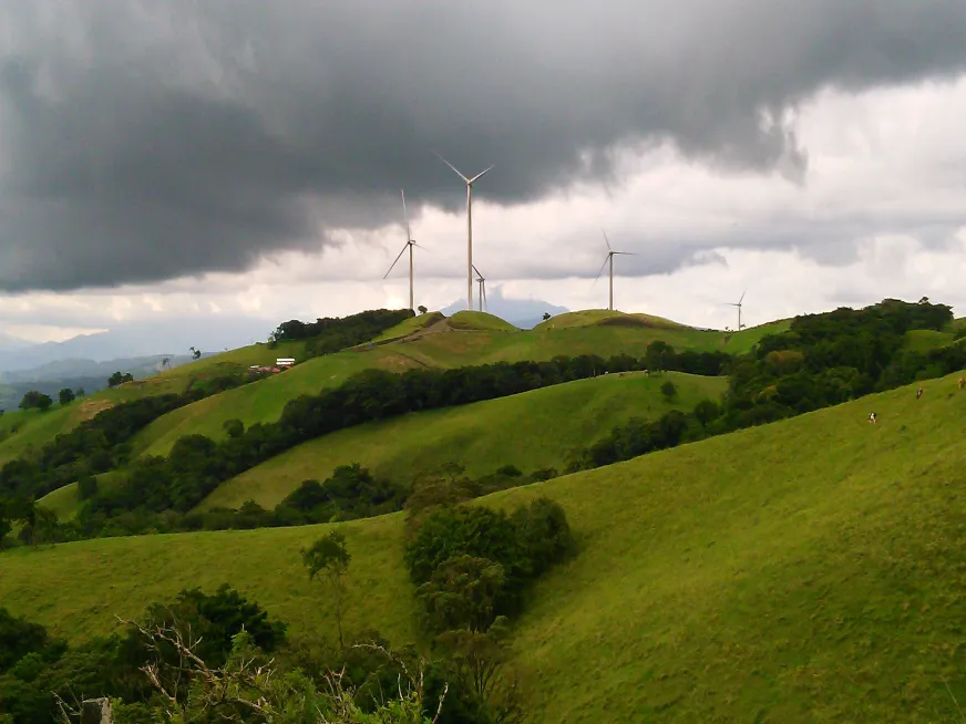 Wind turbines on green hills under a dark, cloudy sky in a rural landscape.