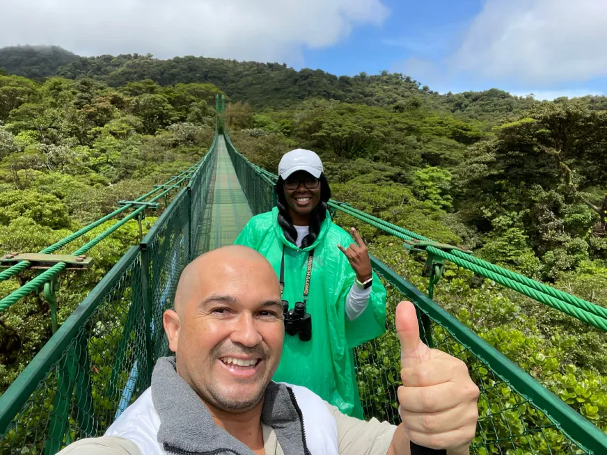 A couple smiles on a suspension bridge surrounded by a lush, green tropical forest.