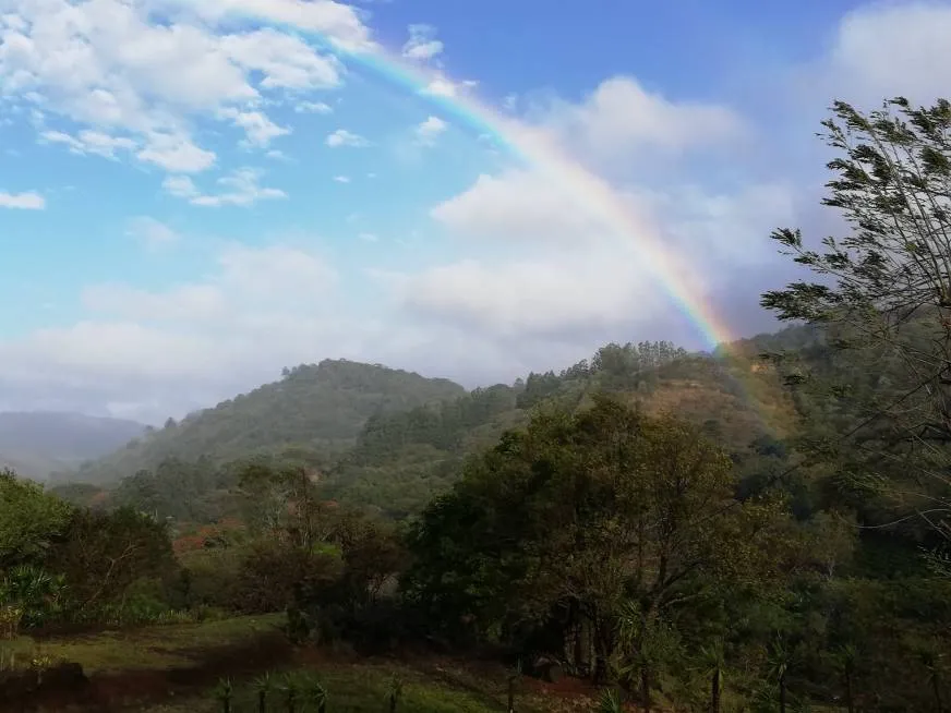 A bright rainbow crosses the sky over green hills covered with trees and white clouds.