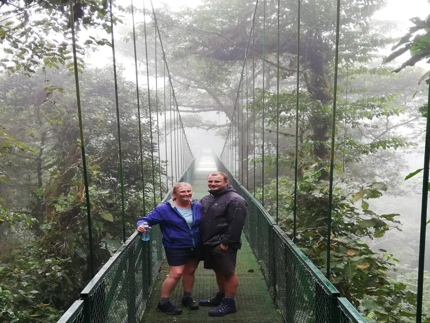 A smiling couple on a suspension bridge surrounded by trees and fog in a tropical forest.