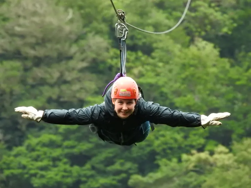 Person wearing an orange helmet and gloves sliding down a zip line against a green forest background.