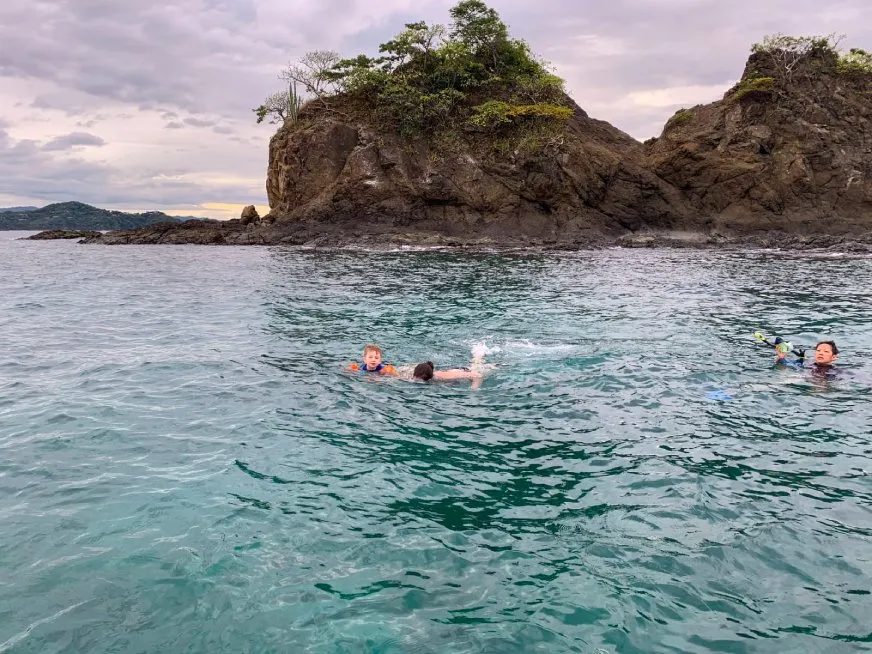 Snorkelers swim in calm blue water near dramatic rocky island formations under a cloudy sky.