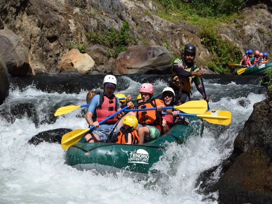 A green raft with tourists paddles through a rocky whitewater canyon surrounded by boulders.