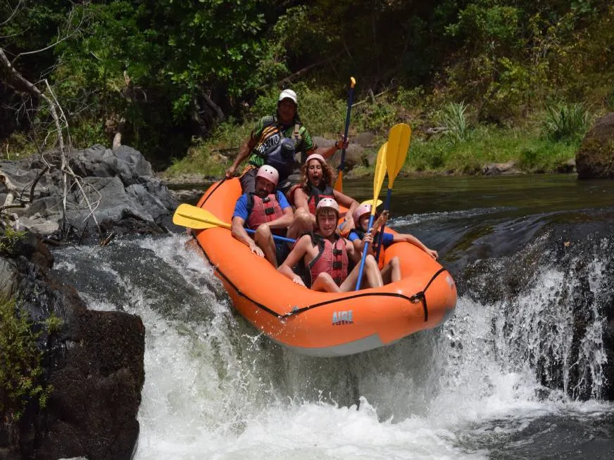 An orange inflatable raft with tourists navigates a powerful whitewater rapid between dark rocky walls.