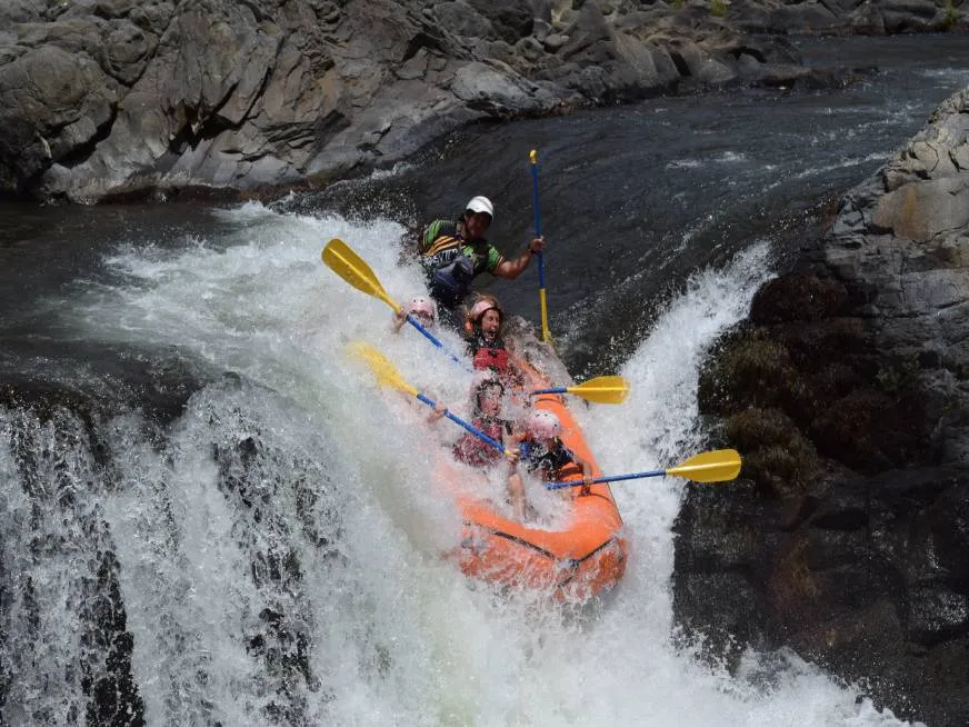 A kayaker in a blue helmet navigates through a narrow whitewater rapid between dark rocky walls.