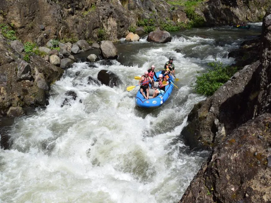 A blue inflatable raft with tourists rushes through a powerful whitewater rapid surrounded by rocks.