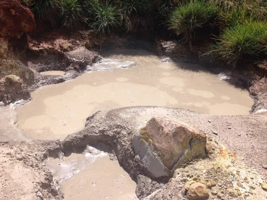 A boiling mud pool bubbles on barren volcanic terrain surrounded by dry rocky ground.