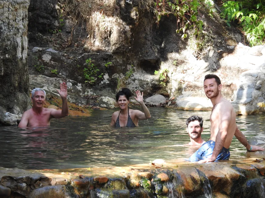 A group of tourists smile while bathing in a natural thermal pool among rocky boulders.