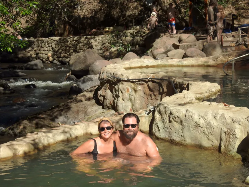 Two tourists smile while soaking in a natural rocky thermal pool surrounded by vegetation.