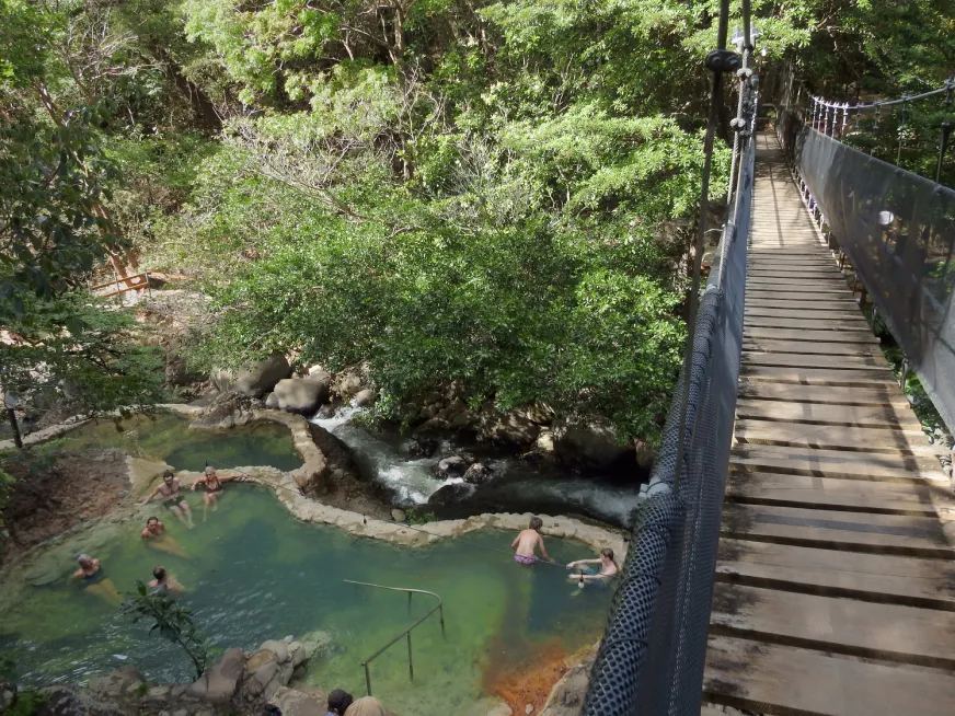 A wooden footbridge crosses a turquoise stream surrounded by lush green tropical vegetation.