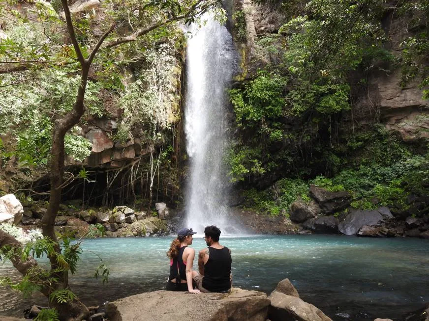 Two hikers stand at the base of a tall waterfall dropping into a jungle pool below.