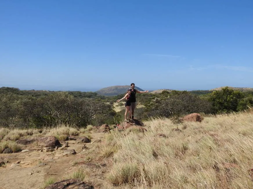 A hiker walks along an open dry trail with a clear blue sky and green hills in the background.