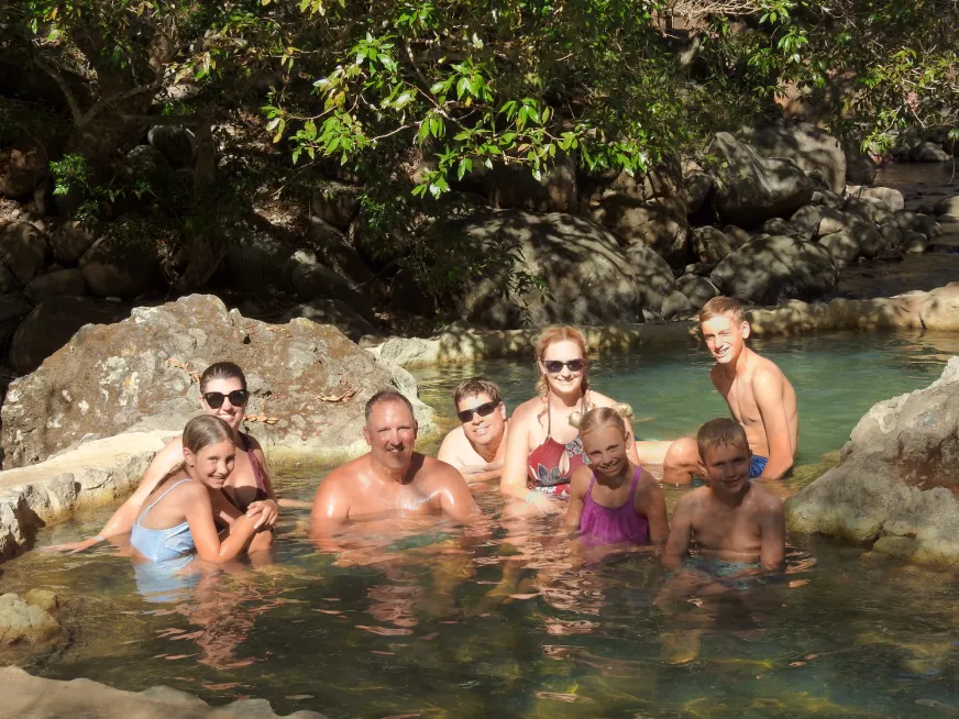 A group of tourists soak and smile in a natural thermal pool surrounded by forest.