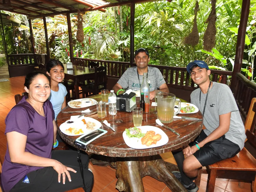 A couple smiles while sharing a meal at a wooden table in an open-air rainforest restaurant.