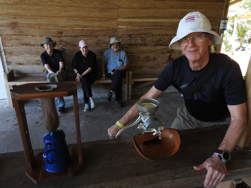 A tourist guide demonstrates a traditional activity to visitors in a rustic open-air building.