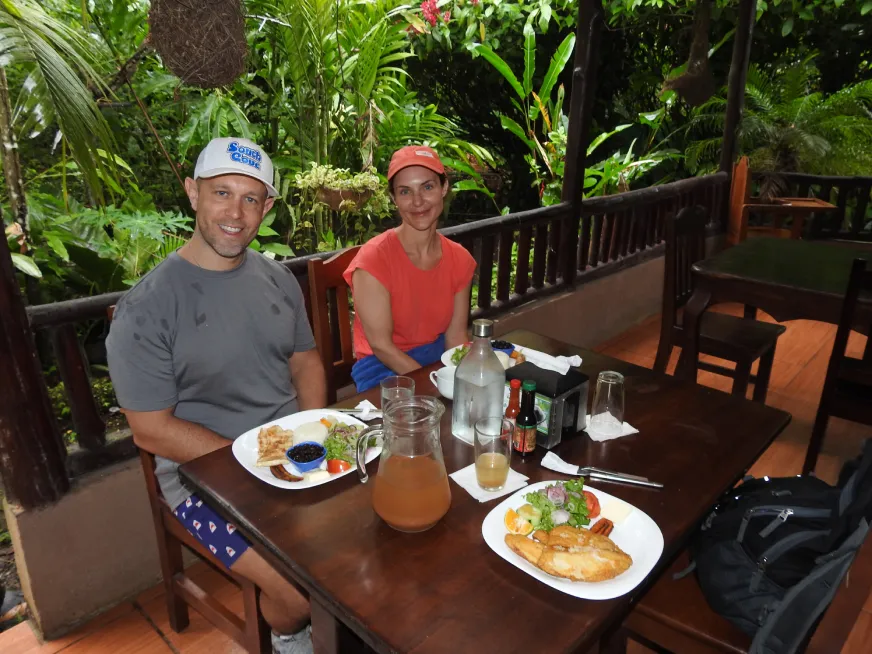 Two tourists smile while having lunch at a wooden table on an open-air jungle terrace.