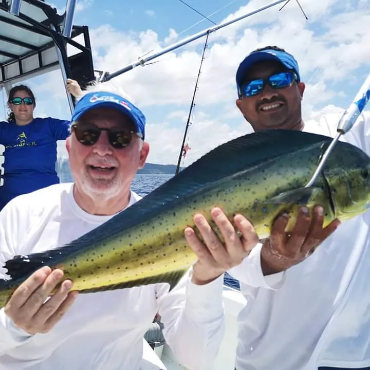 Two smiling men hold a large goldfish on a boat under a sunny sky.