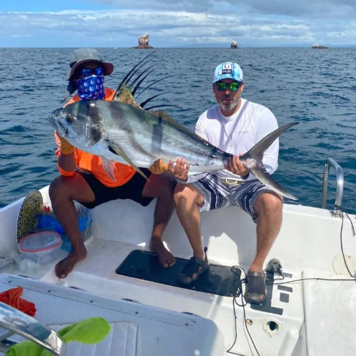 Two men sitting in a boat hold a large roosterfish over the open sea.
