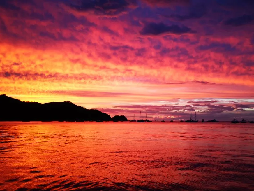 A vivid red and orange sunset sky reflects its colors across a still ocean bay.