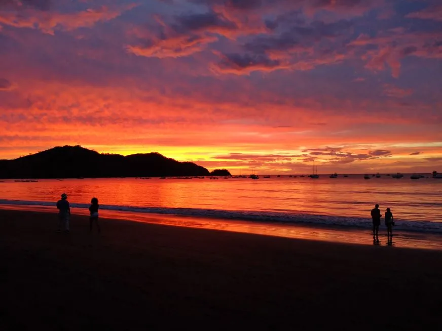 Two people are silhouetted walking on a beach against a deep red and orange sunset sky.