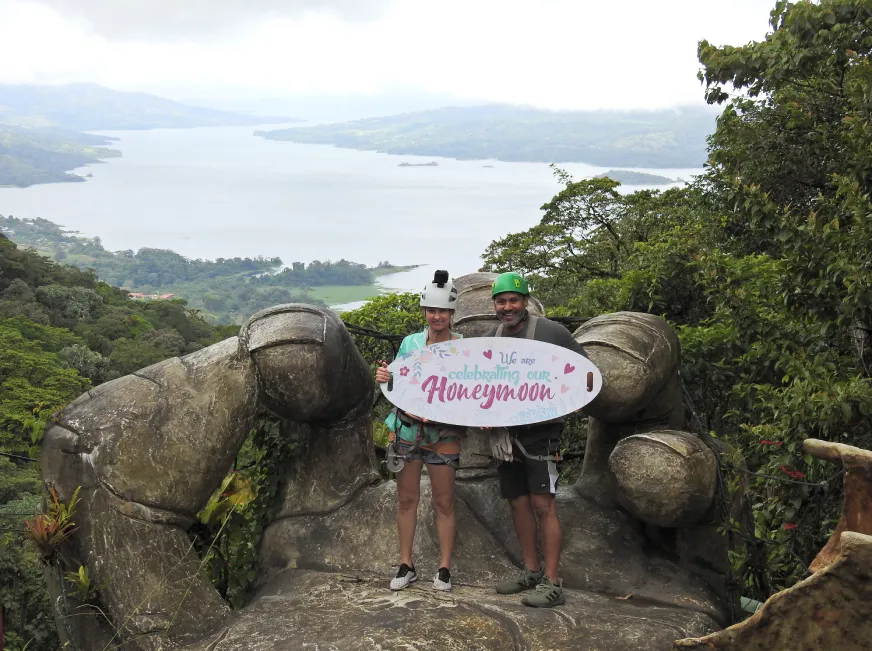 A tourist poses smiling next to a Monteverde sign on a rocky viewpoint surrounded by vegetation.