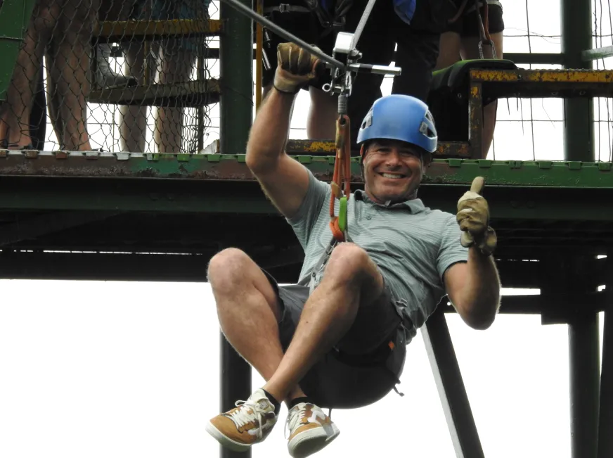 A smiling tourist wearing a blue helmet sits on a zip line platform above a jungle canopy.