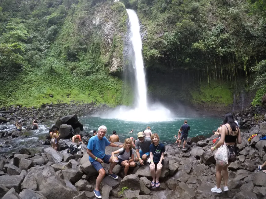 A group of hikers pose smiling in front of a tall waterfall cascading over dark volcanic rocks.